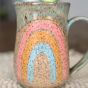 May include: A speckled ceramic mug with a pastel rainbow design. The rainbow features pink, orange, and blue arches. The mug has a light blue handle and rim. The mug is approximately 5 inches tall.