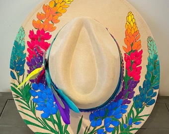 May include: A beige cowboy hat with a painted design of bluebonnets in various shades of blue, purple, pink, orange, and yellow. The flowers are arranged in a circle around the hat.