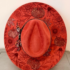 May include: A wide-brimmed orange hat featuring a textured surface with a printed pattern of pumpkins, autumn leaves, and various flowers. The crown is indented, and a small patterned fabric ribbon is attached to the side of the hat brim.