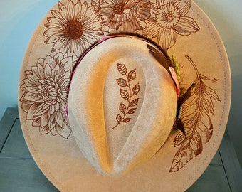 May include: A beige felt fedora hat with brown floral and feather designs. The hat has a wide brim and a crown with a leaf design.