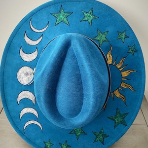 May include: A blue felt fedora hat with a sun and moon design. The hat is decorated with silver moons and a gold sun with flames. There are also green glitter stars around the brim.