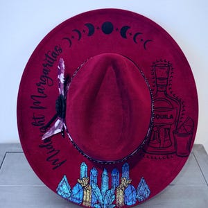 May include: A deep red wide-brimmed hat featuring black moon phase graphics, the text Midnight Margaritas, a tequila bottle and glass illustration, and a cluster of blue and gold crystals. A patterned fabric ribbon is tied around the crown.