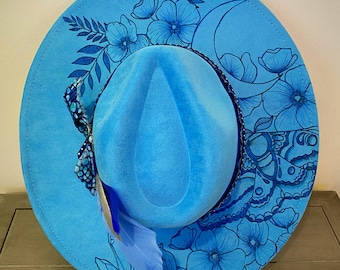 May include: A light blue fedora hat with a wide brim, decorated with blue floral and moon designs. A blue feather and patterned ribbon adorn the side. The hat is a stylish accessory.