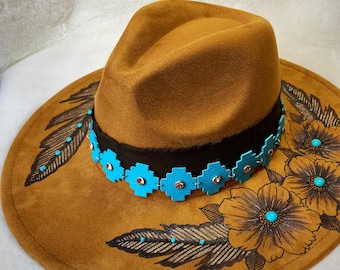 May include: A brown fedora-style hat with a wide brim. The brim is decorated with etched floral and feather designs, accented with turquoise-colored embellishments. A black band encircles the base of the crown, adorned with turquoise floral accents and small orange gems.