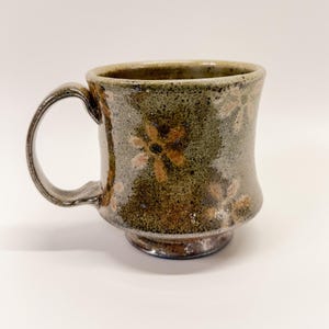 May include: A handmade ceramic mug with a dark green and brown glaze. The mug features a floral design in shades of orange and brown. It has a rounded base and a sturdy handle.