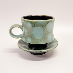 May include: A ceramic teacup and saucer set. The cup and saucer are a speckled seafoam green with large, light blue polka dots. The interior of the cup is dark brown. The set is on a white background.