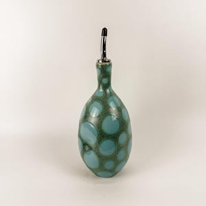 May include: A teal and green ceramic oil bottle with a stainless steel spout. The bottle has a rounded shape and is decorated with large, irregular teal-colored polka dots against a darker green background. The spout has a black tip.