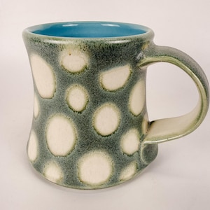 May include: A ceramic mug with a curved handle, featuring a textured exterior with a mottled dark green and grey glaze over a series of circular white spots. The interior of the mug is finished in a smooth, solid light blue glaze.