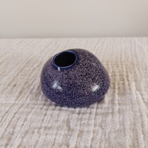 Puede incluir: Un peque&ntilde;o jarr&oacute;n de cer&aacute;mica redondeado con un acabado moteado en morado y azul marino. El jarr&oacute;n tiene una sola abertura en la parte superior y una forma suave, similar a un guijarro. El interior de la abertura es azul marino oscuro.