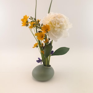 May include: A small, green ceramic vase with a rounded shape holds a bouquet of white peonies and yellow flowers.