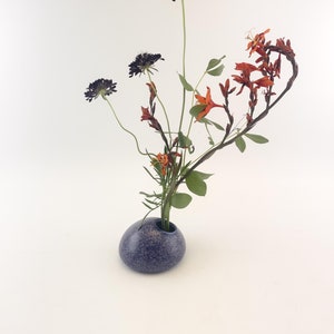 Puede incluir: Un peque&ntilde;o jarr&oacute;n azul moteado contiene un arreglo floral minimalista. El arreglo presenta flores oscuras y esf&eacute;ricas, flores naranjas y hojas verdes. El jarr&oacute;n tiene forma redondeada y una abertura estrecha, sobre un fondo blanco.