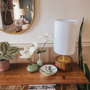 Puede incluir: Una consola de madera con una l&aacute;mpara blanca, lirios blancos en peque&ntilde;os jarrones y un plato decorativo. Una planta en maceta y una planta de serpiente est&aacute;n a ambos lados. Un espejo refleja una sala de estar.