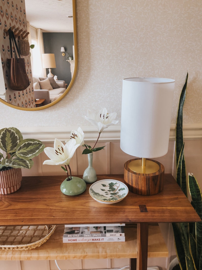 Puede incluir: Una consola de madera con una l&aacute;mpara blanca, lirios blancos en peque&ntilde;os jarrones y un plato decorativo. Una planta en maceta y una planta de serpiente est&aacute;n a ambos lados. Un espejo refleja una sala de estar.