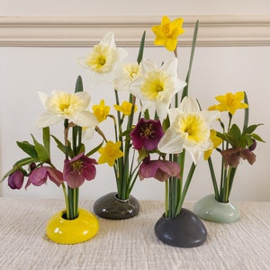 Puede incluir: Arreglo floral con narcisos blancos y amarillos, y flores de helleborus moradas oscuras. Las flores se exhiben en peque&ntilde;os jarrones en forma de guijarro en amarillo, gris y verde. El arreglo est&aacute; sobre una superficie texturizada de color neutro.