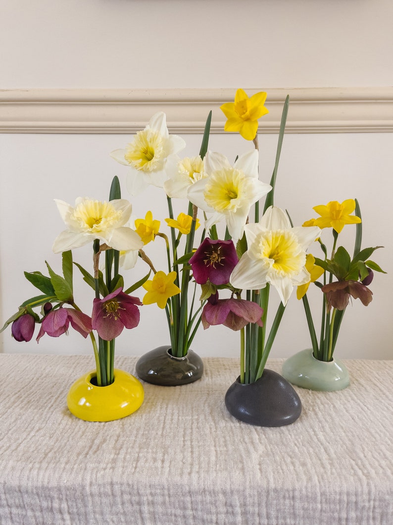 Puede incluir: Arreglo floral con narcisos blancos y amarillos, y flores de helleborus moradas oscuras. Las flores se exhiben en peque&ntilde;os jarrones en forma de guijarro en amarillo, gris y verde. El arreglo est&aacute; sobre una superficie texturizada de color neutro.