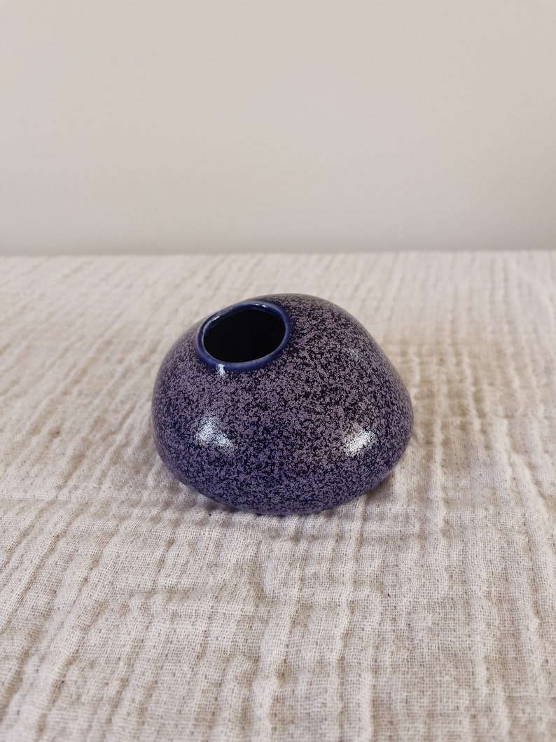 Puede incluir: Un peque&ntilde;o jarr&oacute;n de cer&aacute;mica redondeado con un acabado moteado en morado y azul marino. El jarr&oacute;n tiene una sola abertura en la parte superior y una forma suave, similar a un guijarro. El interior de la abertura es azul marino oscuro.