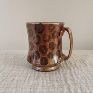May include: A brown ceramic mug with a speckled pattern of brown and black circles. The mug has a flared base and a rounded handle.