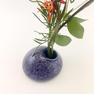 Puede incluir: Un peque&ntilde;o jarr&oacute;n azul moteado de forma redonda contiene un arreglo floral. El jarr&oacute;n tiene una abertura azul oscuro y una superficie texturizada. El arreglo incluye hojas y tallos verdes, con flores naranjas y amarillas.