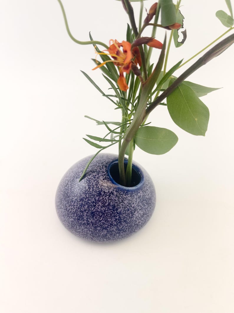 Puede incluir: Un peque&ntilde;o jarr&oacute;n azul moteado de forma redonda contiene un arreglo floral. El jarr&oacute;n tiene una abertura azul oscuro y una superficie texturizada. El arreglo incluye hojas y tallos verdes, con flores naranjas y amarillas.