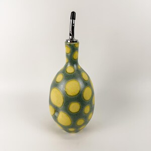 May include: A ceramic olive oil bottle with a green base and large yellow polka dots. The bottle has a long, slender neck and a stainless steel spout with a black tip. The bottle is designed for dispensing oil or vinegar.