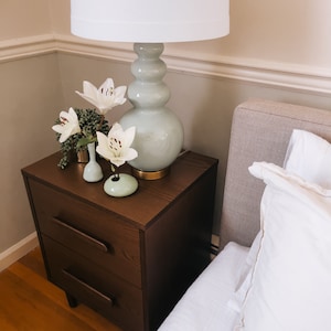 Puede incluir: Una escena de dormitorio con una mesita de noche de madera marr&oacute;n oscuro con dos cajones, coronada por una l&aacute;mpara verde claro y una pantalla blanca. Tambi&eacute;n se ven lirios blancos en peque&ntilde;os jarrones y una cama con ropa de cama blanca.