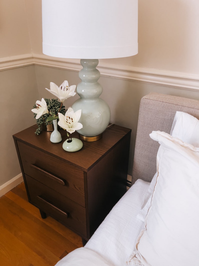 Puede incluir: Una escena de dormitorio con una mesita de noche de madera marr&oacute;n oscuro con dos cajones, coronada por una l&aacute;mpara verde claro y una pantalla blanca. Tambi&eacute;n se ven lirios blancos en peque&ntilde;os jarrones y una cama con ropa de cama blanca.