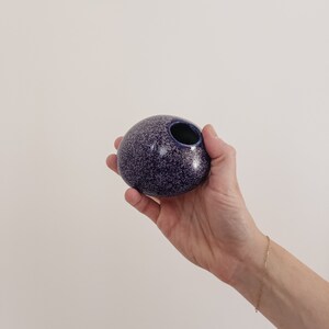 Puede incluir: Un peque&ntilde;o jarr&oacute;n redondo de cer&aacute;mica morada moteada con interior y abertura azul oscuro. El jarr&oacute;n se sostiene en una mano sobre un fondo neutro. Es un objeto decorativo.
