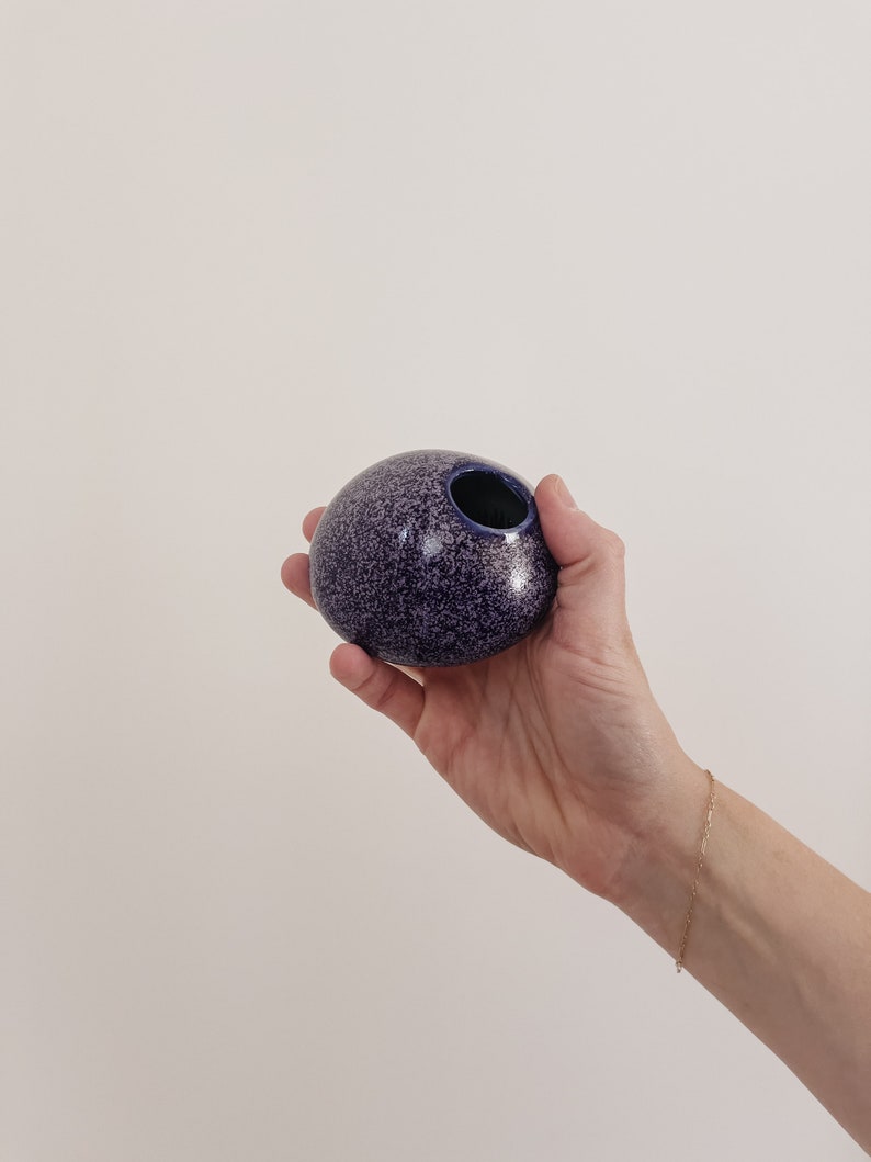 Puede incluir: Un peque&ntilde;o jarr&oacute;n redondo de cer&aacute;mica morada moteada con interior y abertura azul oscuro. El jarr&oacute;n se sostiene en una mano sobre un fondo neutro. Es un objeto decorativo.