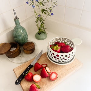 Puede incluir: Una escena de cocina con fresas frescas, una tabla de cortar de madera y un cuchillo. Un cuenco de cer&aacute;mica contiene fresas enteras, mientras que las cortadas descansan sobre la tabla. Tambi&eacute;n hay una botella de aceite verde y un jarr&oacute;n con flores azules.