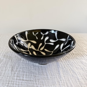 May include: A small, round black ceramic bowl with a glossy finish. The bowl features a white, leaf-like pattern. The bowl is likely used for serving or display.