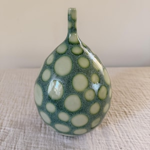 May include: A handcrafted ceramic vase with a unique, organic shape. The vase features a speckled teal background with large, irregular, light green polka dots. The vase has a narrow neck and a bulbous body, perfect for displaying flowers.
