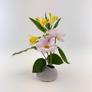 May include: A small, gray ceramic vase holds a floral arrangement. The arrangement features light pink daisies, yellow lilies, and green leaves. The vase has a rounded shape and a narrow opening, creating a minimalist aesthetic.
