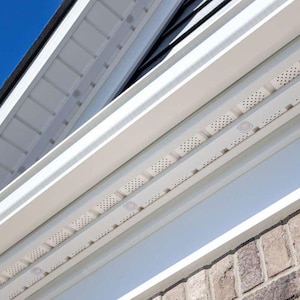 Puede incluir: Vista exterior de una casa con molduras y revestimientos blancos. La imagen muestra una sección de la línea del techo, con paneles de alero y tablas de fascia blancas. El revestimiento es de color gris claro y la mampostería es visible en la parte inferior.