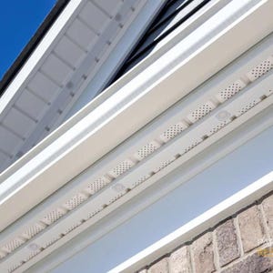 May include: A close-up shot of white soffit and trim against a brick wall. The soffit features small, regularly spaced perforations. A clear blue sky is visible. The image is well-lit, showcasing the architectural design.