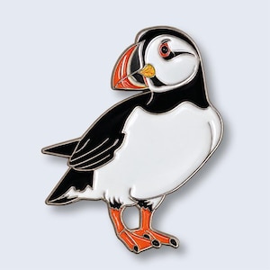 May include: A metal enamel pin shaped like a puffin. The bird features a black head and back, a white chest and belly, and a distinctive beak with orange, yellow, and black markings. The puffin has bright orange webbed feet and a red eye.