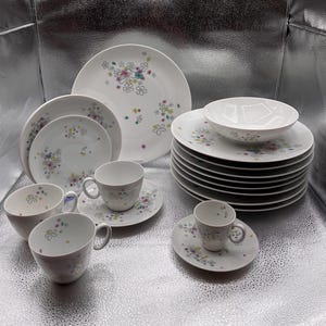 May include: A white porcelain dinnerware set featuring a floral pattern in pastel colors. The set includes plates, bowls, cups, and saucers. The floral design is composed of small, colorful flower clusters. The dishes are arranged on a reflective surface.