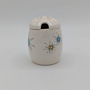 May include: A speckled, off-white ceramic sugar bowl with a lid. The bowl features a mid-century modern design with blue and yellow starburst patterns. The lid has a slot for a spoon.