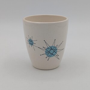 May include: A cream-colored ceramic tumbler with a speckled finish. The tumbler features two blue starburst designs with black lines. The interior of the tumbler is a light beige color. The design is reminiscent of mid-century modern aesthetics.