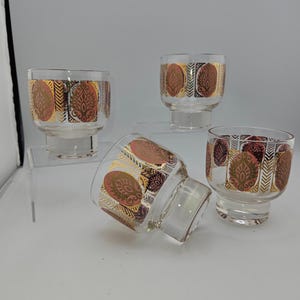 May include: Four clear glass tumblers with a decorative gold, red, and white pattern. The pattern features an oval design with a gold border and a repeating arrow design. The glasses are on a clear surface.