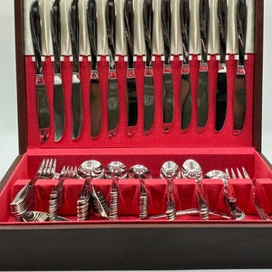 May include: A set of stainless steel flatware including knives, forks, and spoons arranged in a dark wooden storage box with a red velvet interior. The knife handles feature a decorative star pattern and a curved, polished metallic finish.