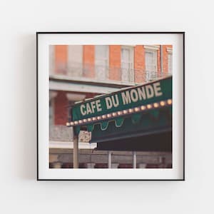 May include: Square framed print featuring the Cafe Du Monde sign in New Orleans. The sign is green with white lettering and small lights. The background shows a brick building with white windows and a wrought iron balcony.