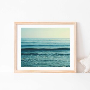 May include: A framed photograph of a blue ocean with waves crashing on the shore. The photo is in a light wood frame and is hanging on a white wall.