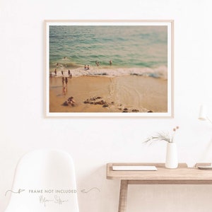 May include: A blurry photograph of a beach scene with people swimming in the ocean. The water is a light blue color and the sand is a light brown color. The photo has a vintage filter applied to it.