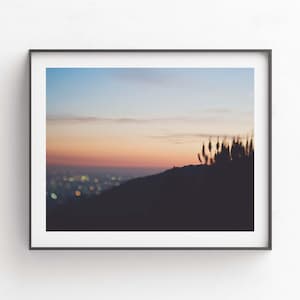 Op de afbeelding: Een ingelijste foto van een zonsondergang boven een stadsbeeld. De stadslichten zijn wazig in de verte, en de lucht is zacht roze en oranje. Silhouetten van bomen en gras bevinden zich op de voorgrond.