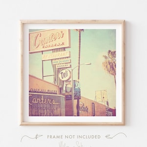 May include: A vintage photograph of a retro diner called "Canters" in Fairfax, California. The sign is lit up with neon lights and the building is painted in pastel colours. The photo has a faded, vintage look.