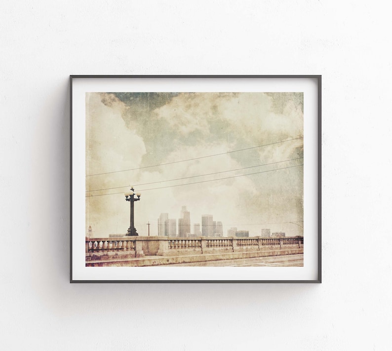 May include: A vintage-style photograph of a cityscape with a bridge and a streetlight in the foreground. The sky is cloudy and the city skyline is visible in the distance.