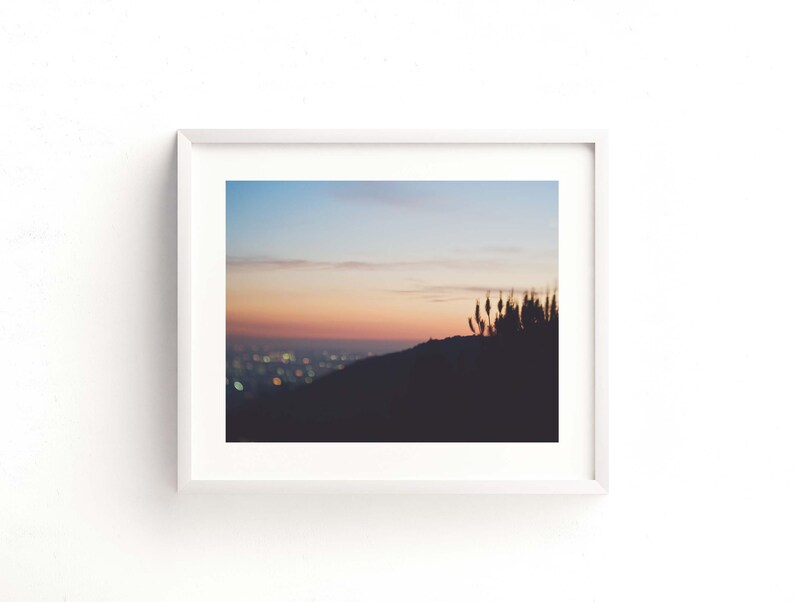 Op de afbeelding: Een ingelijste foto van een zonsondergang boven een stadsbeeld. De lucht is een gradi&euml;nt van roze, oranje en blauw, met een paar wolken. De stadslichten zijn in de verte zichtbaar, en er zijn wat hoge grassen op de voorgrond.