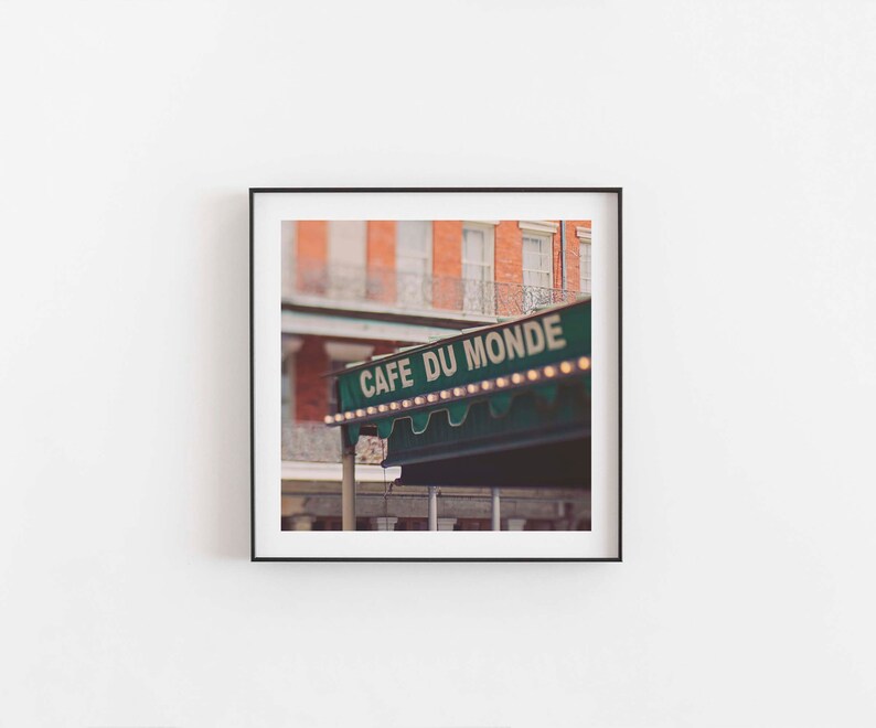 May include: Square framed print featuring the Cafe Du Monde sign in New Orleans. The sign is green with white lettering and small lights. The background shows a brick building with white windows and a wrought iron balcony.