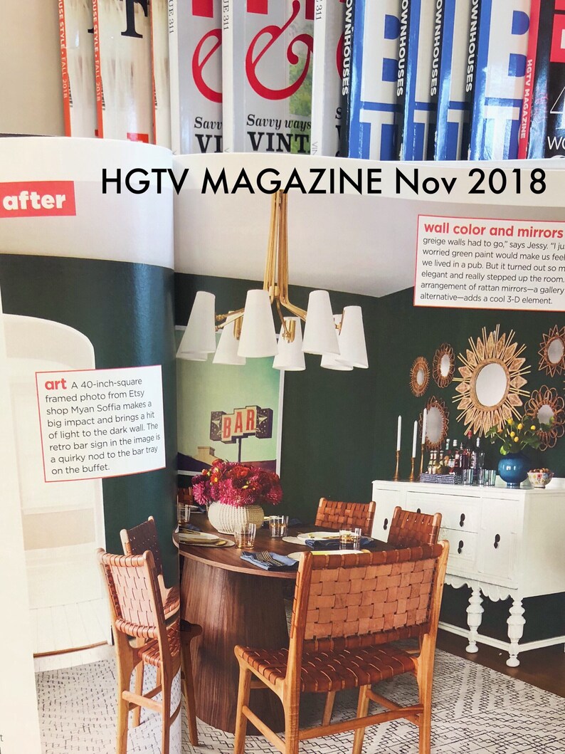 May include: A dining room with a dark green wall, a white buffet, and a wooden table with four chairs. The chairs are made of woven leather and have a natural brown color. The table is round and has a dark wood finish. The buffet has a white finish and has a white countertop. The wall is decorated with a gallery of round mirrors and a large framed photo of a bar sign. The photo is 40 inches square and is framed in a black frame. The mirrors are all different sizes and shapes and are made of rattan. The room is decorated in a mid-century modern style.
