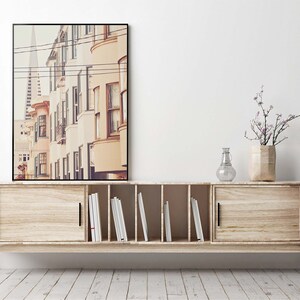 May include: A light wood cabinet with two sliding doors and a center shelf with books. A framed photograph of a city street with buildings and power lines hangs above the cabinet. A vase with a small bouquet of purple flowers sits on the cabinet.